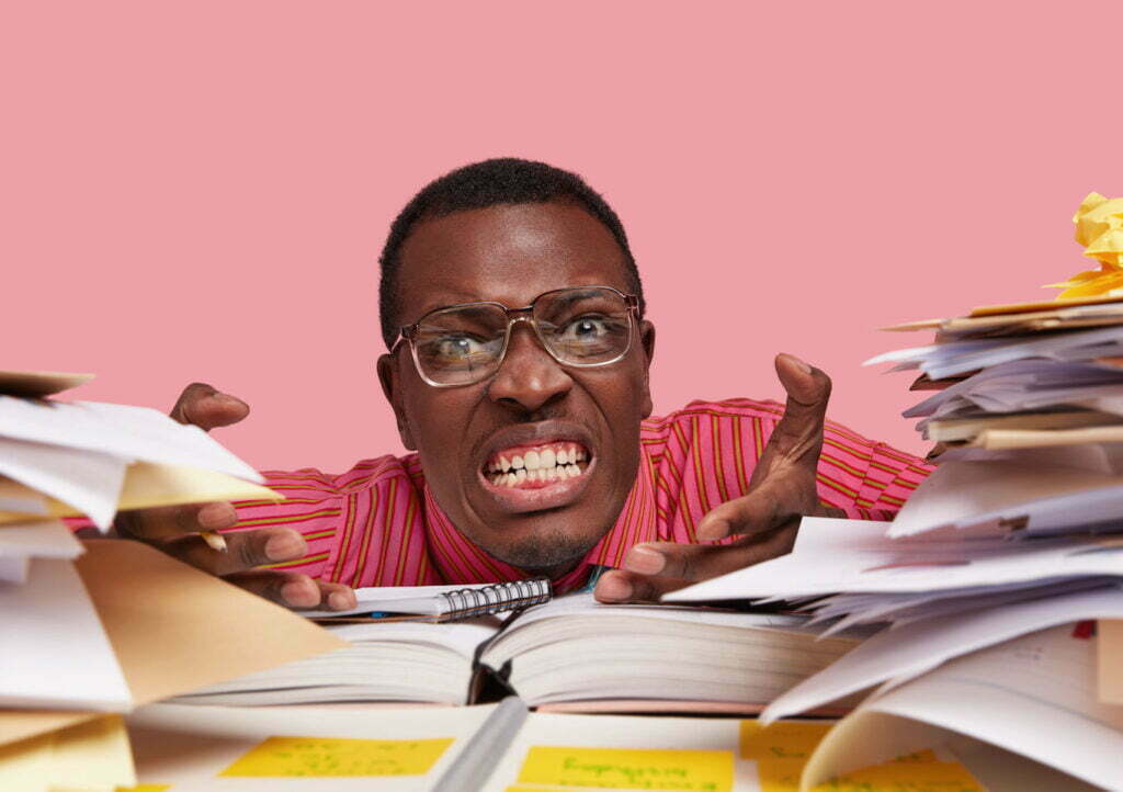 Stressful annoyed tired teacher clenches teeth in anger, gestures and looks directly at camera, feels fatigue of preparing for lessons or making project, surrounded with papers, being overload my story of perfectionism