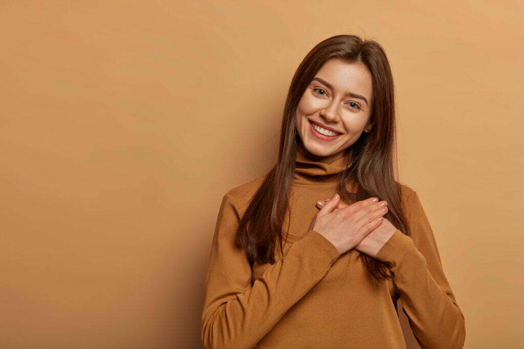 Moment of love and appreciation. Adorable cheerful woman keeps both palms pressed together near heart in gratitude symbol, smiles gently, thanks fate for wellbeing, expresses sincere emotions You matter
